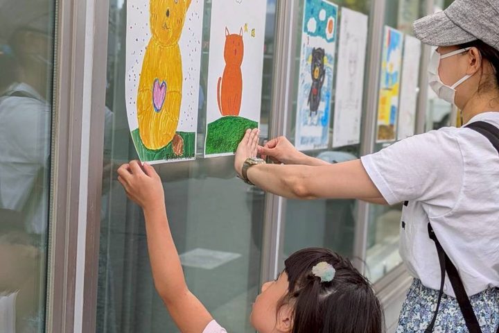 子どもが描いた絵をお店の窓に貼るのを、スタッフが手伝っているようす